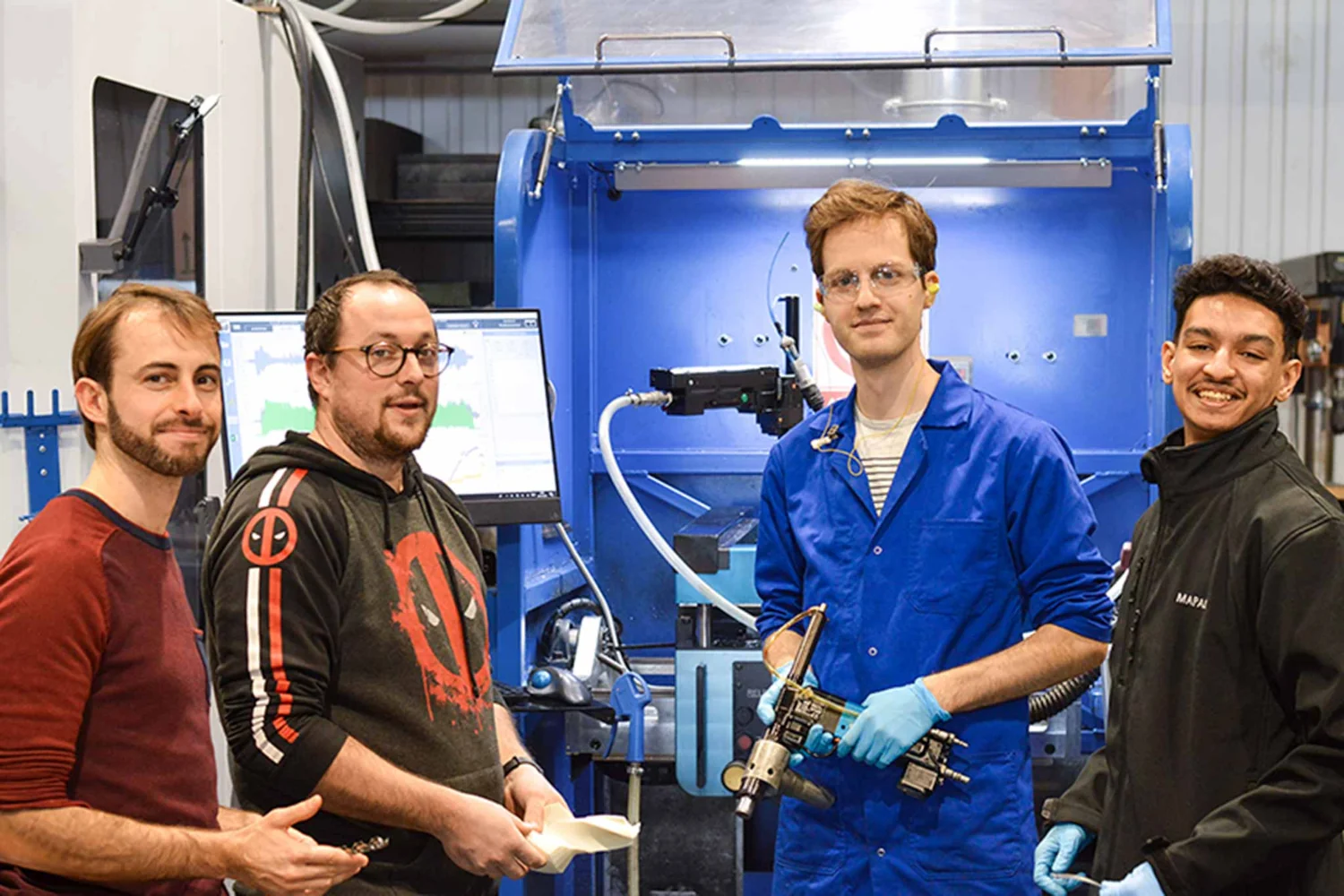 Four men stand in a manufacturing environment. One has a hand drill in his hand.